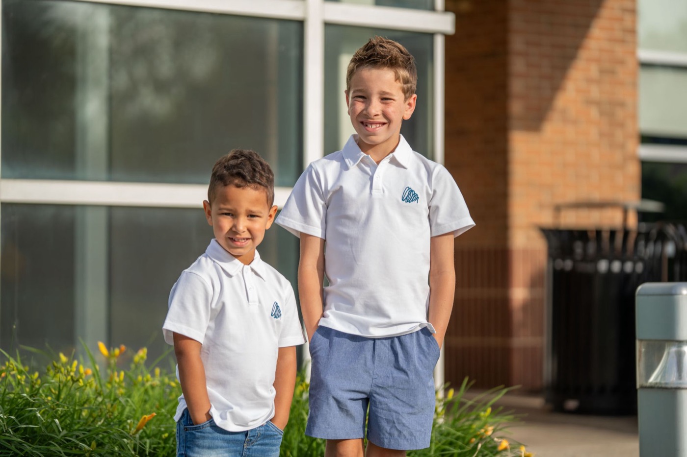 Two smiling boys in white Cam & Coop polos standing together in front of a brick building